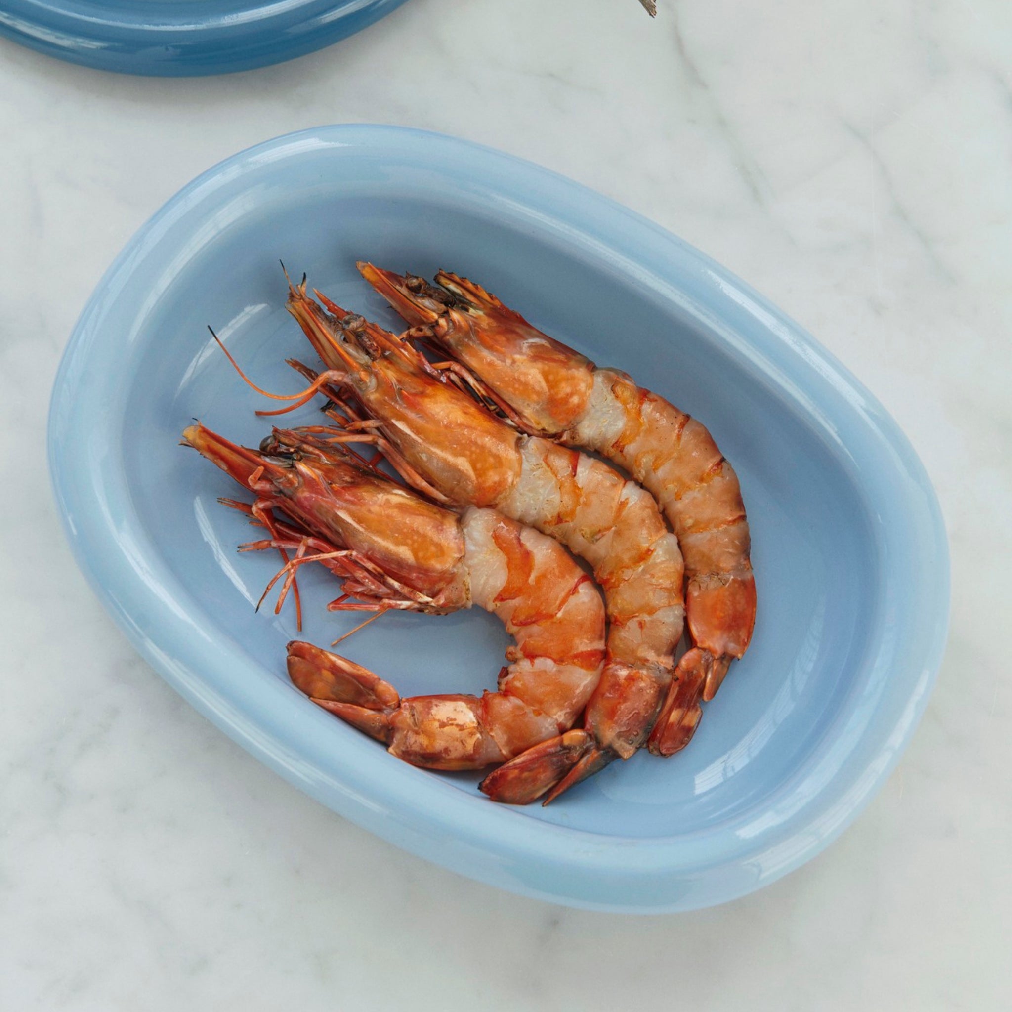 Vier gekookte garnalen staan in een gebogen lijn op de Hay Barro Ovale Schaal Lichtblauw 20/27 van Hay, geplaatst op een wit marmeren oppervlak.