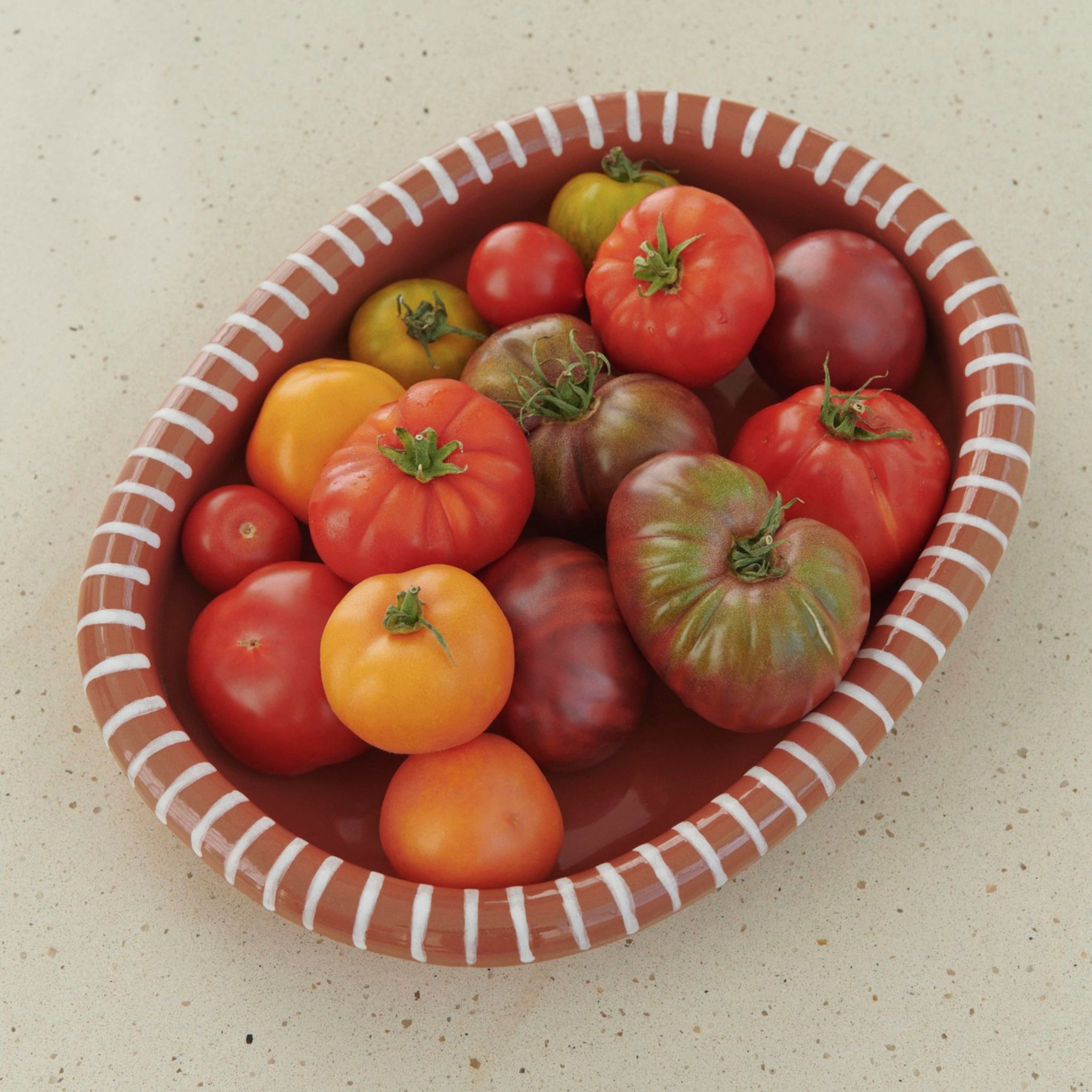De Hay Barro Ovale Schaal Terracotta 27/36 van Hay heeft witte accenten en een mix van levendige heirloom tomaten - rood, oranje, groen en paars - op het licht gespikkelde oppervlak.