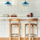 Een moderne keuken met twee houten barkrukken op een wit betegeld aanrecht, gestapelde borden, kopjes en keukengerei, waaronder de Hay Barro Ovale Schaal Terracotta 20/27 van Hay, met twee blauwe hanglampen boven je hoofd.