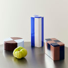 Drie containers van Hay Blikken Doos - in blauw, gebroken wit en bruin - hebben ronde, hoge rechthoekige en korte rechthoekige vormen op een grijs oppervlak met een groene heirloom tomaat ervoor.