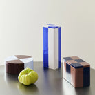 De Hay Blikken Doos Bordeaux Ijsblauw Rond op een donkere tafel naast een groene tomaat, met een blauw-witte en een geblokte blikken doos.