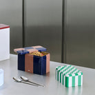 Een open rechthoekig blik met een roze en marineblauw geruit deksel onthult ontbijtgranen, terwijl de Hay Blikken Doos Groen Off-White S van Hay, met een groen dambordmotief, vlakbij staat. Twee lepels rusten op het metalen aanrecht.