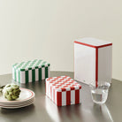 De Hay Blikken Doos Rood Off-White S met rood-witte strepen op een ronde tafel, naast een groen-witte blikken doos, een hoge blikken doos en een artisjok.