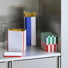 De Hay Blikken Doos Groen Off-White S van Hay voegt een stijlvolle opbergoplossing toe aan een metalen aanrecht. Het houdt pasta vast te midden van levendige rode, witte, blauwe en groene patronen met een glas water in de buurt.