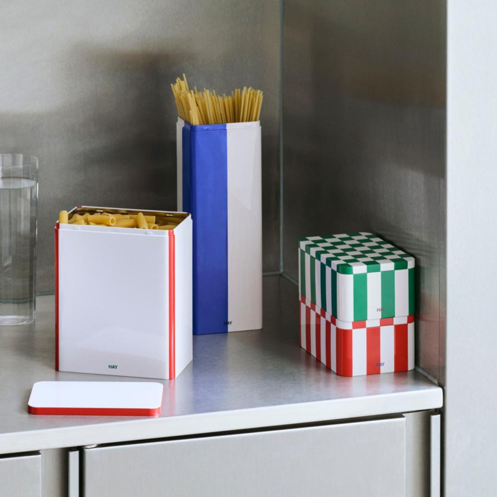 De Hay Blikken Doos Groen Off-White S van Hay voegt een stijlvolle opbergoplossing toe aan een metalen aanrecht. Het houdt pasta vast te midden van levendige rode, witte, blauwe en groene patronen met een glas water in de buurt.