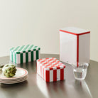 De Hay Blikken Doos Groen Off-White S van Hay voegt charme toe als keukenaccessoire, in combinatie met andere geruite en gestreepte blikken op een ronde tafel met borden, een artisjok en een glas water.