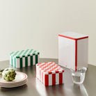 Drie Hay Blikken Doos Off-White Rood L containers met dambordmotieven, twee rood-wit en een groen-wit, staan op een tafel met borden, een artisjok en een glas water tegen een neutrale achtergrond.