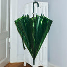 De Hay Canopy Paraplu Groen van het merk Hay, gemaakt van gerecycled materiaal en voorzien van een gebogen handvat, staat gesloten tegen een witte radiator in de hoek van een lichte kamer met houten vloer.