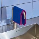 Een Hay Canteen Vaatdoek Blauw Geel van Hay is gedrapeerd over een zilveren kraan boven een roestvrijstalen gootsteen, tegen een lichtblauw betegelde muur - ideaal voor efficiënt keuken schoonmaken.