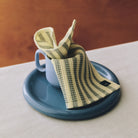 De gestreepte geel-groene Hay Canteen Vaatdoek uit de set Geel Groen hangt over een blauwe mok op een bord.