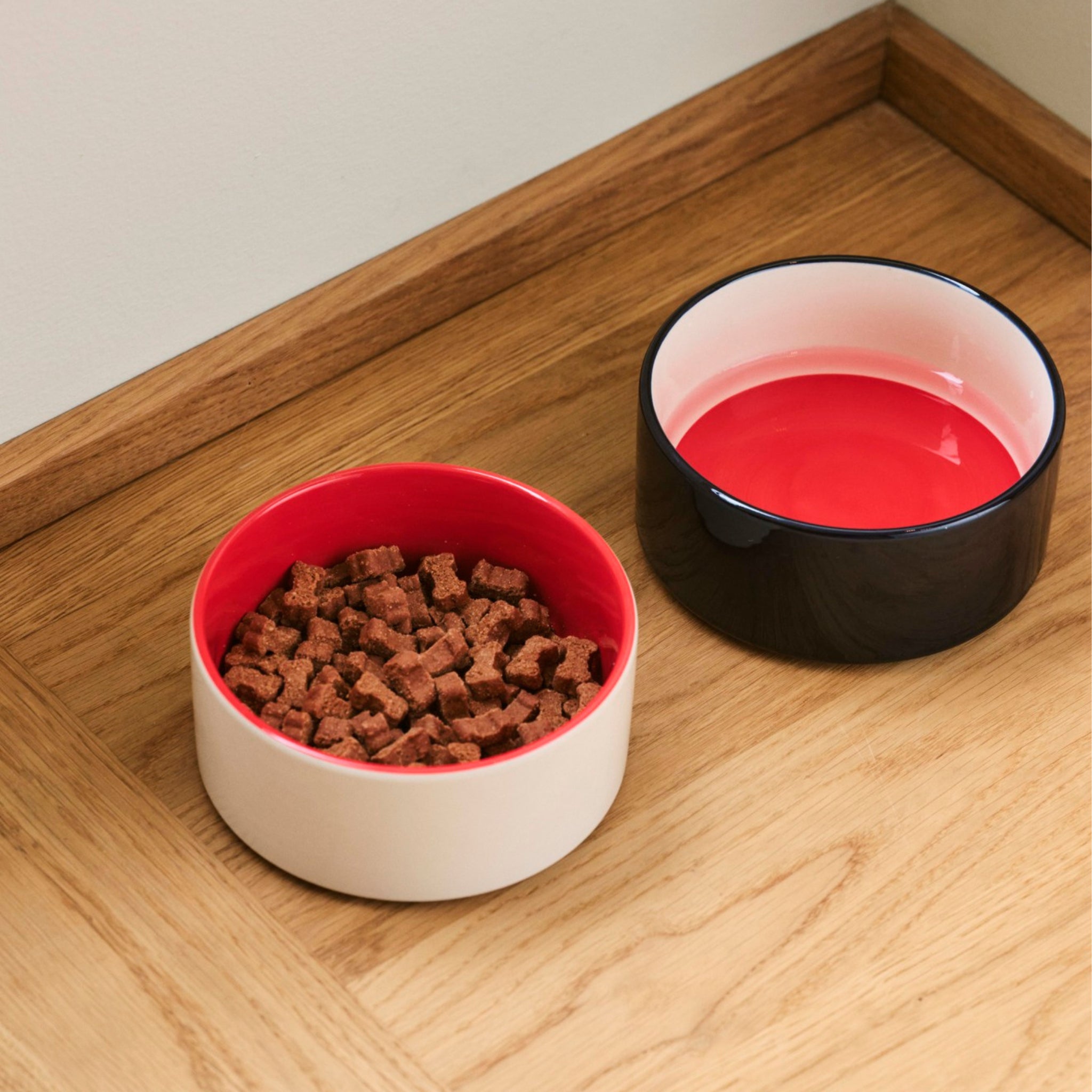 In de hoek op een houten vloer staan twee voerbakken: een Hay Dogs Hondenbak Blauw Rood D15 van Hay, met een rode binnenkant gevuld met brokjes, en een zwarte bak met een rode binnenkant met water.