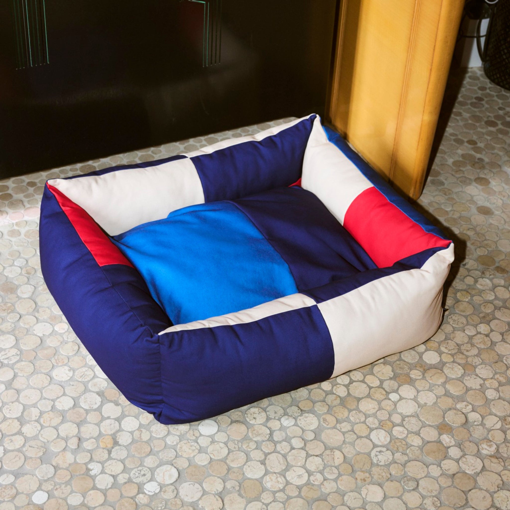 Een vierkante Hay Hondenmand Rood Blauw 65/78 met blauwe, rode en witte patchwork zijkanten ligt op een tegelvloer met kiezelpatroon bij een muur en een houten kast - perfect voor huisdieren die van de gezellige Hay-stijl houden.
