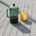 De Hay French Press Koffiemaker Aqua met koffie en een gele mok op een tafel in strepen zonlicht.