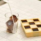 In een glas met koffie-ijsblokjes is melk gegoten. Ernaast staat een Hay ijsblokjesvorm Vierkant XL Blauw van Hay, gevuld met koffie- en melkijsblokjes, alles op een wit betegeld oppervlak.