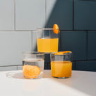 Drie glazen Glas Helder M van Hay staan op een wit oppervlak in het zonlicht: in de ene zit water met een gepelde sinaasappel, in de andere sinaasappelsap met een schijfje sinaasappel en in de derde zit sap met een sinaasappelschil. Blauwe tegelmuur op achtergrond.