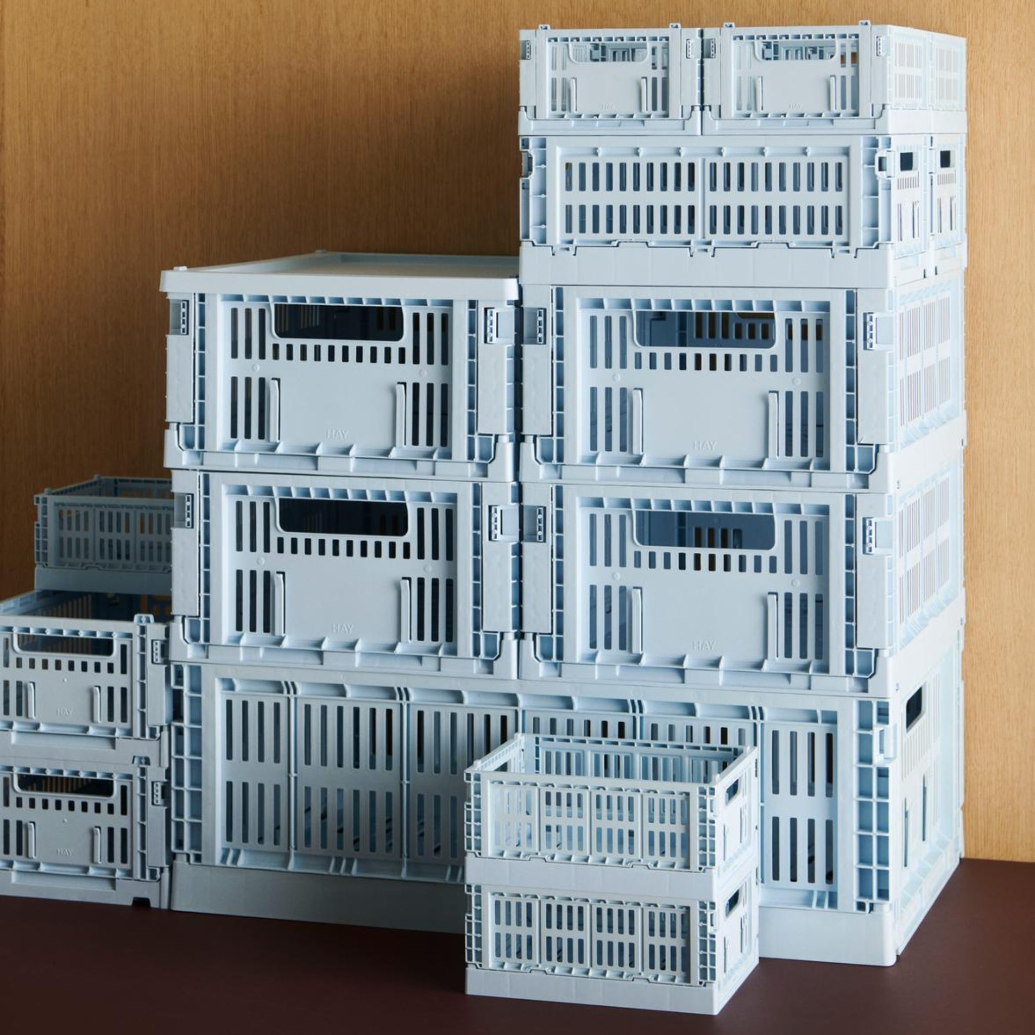 Een stapeltje Hay Kleur Krat Mini Lichtblauw opbergkratten van gerecycled plastic van het merk Hay staan in verschillende maten op een donkerbruin oppervlak tegen een houten achtergrond.