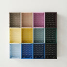 Twaalf lege Hay Kleur Krat Mini Lichtblauw opbergkratten van Hay, gemaakt van gerecycled plastic, in een raster van drie rijen en vier kolommen, zijn gemonteerd op een effen witte muur.