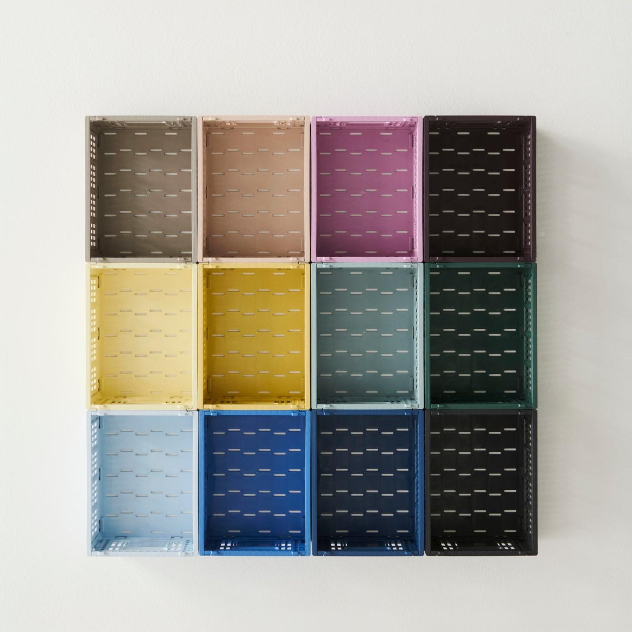 Twaalf lege Hay Kleur Krat Mini Lichtblauw opbergkratten van Hay, gemaakt van gerecycled plastic, in een raster van drie rijen en vier kolommen, zijn gemonteerd op een effen witte muur.