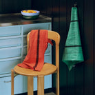 Een Theedoek Rood van Hay Linear is over een houten stoel gedrapeerd, terwijl een groene handdoek aan een haakje in de keuken hangt, met sinaasappels in een schaal op het roestvrijstalen aanrecht erachter.