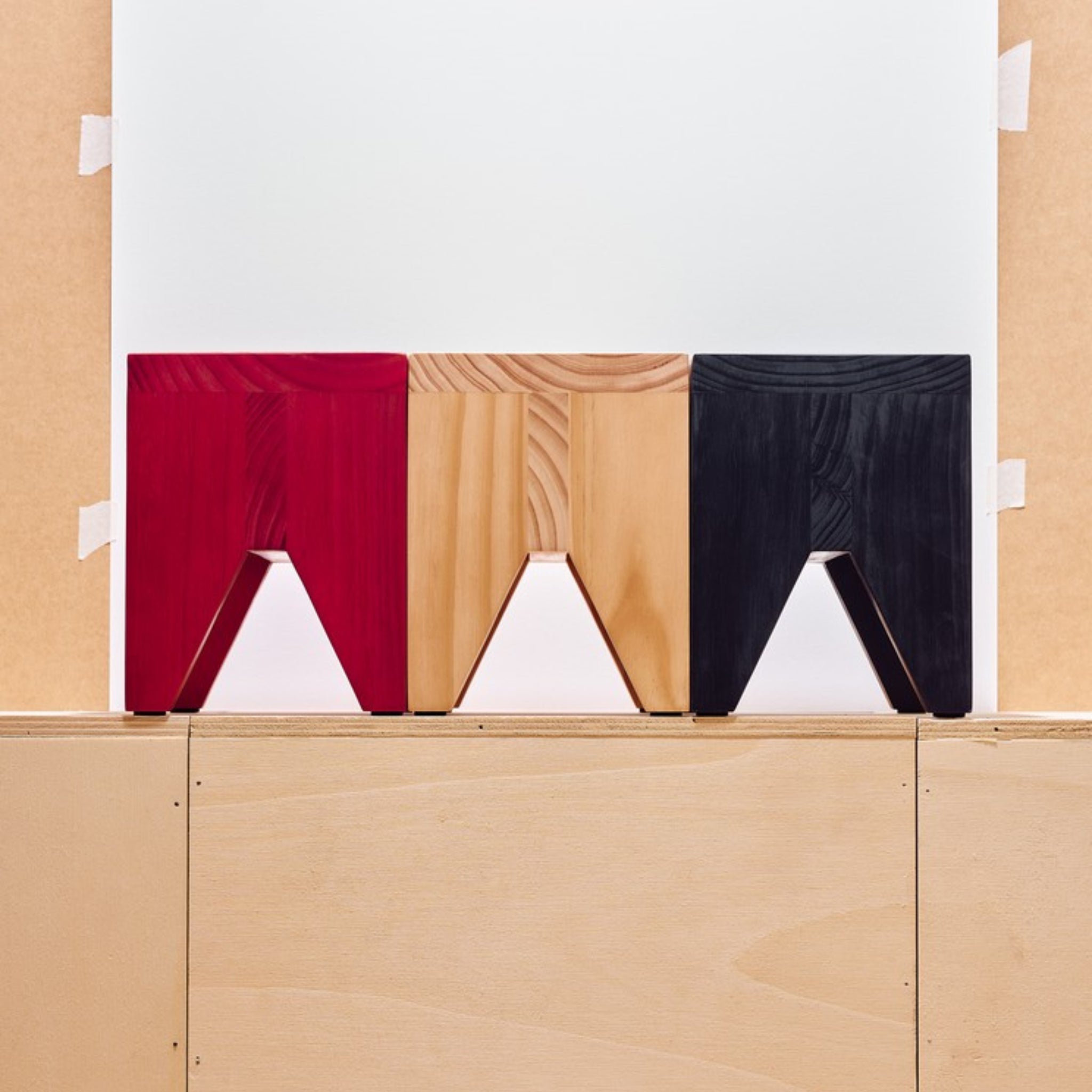 Drie Hay Manolito Kruk Zwart Kort krukken van Hay - rood, naturel hout en zwart - staan naast elkaar tegen een neutrale achtergrond, waarbij hun moderne V-vormige uitsnede benadrukt wordt.