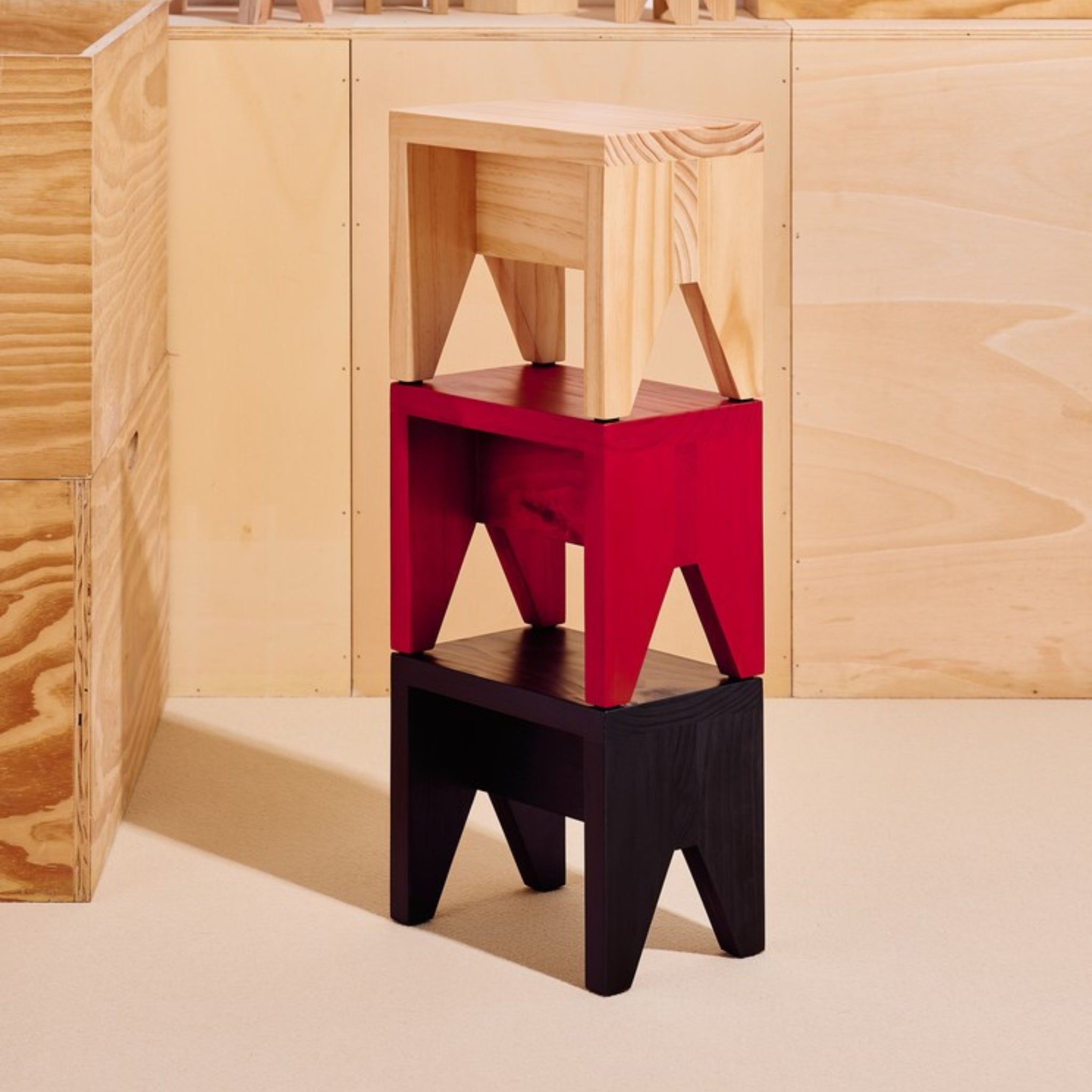 Drie houten krukjes zijn gestapeld: een licht houten exemplaar bovenop, een rood krukje in het midden en de Hay Manolito Kruk Zwart Kort van Hay onderaan. Deze display laat moderne designtrends zien tegen een lichthouten achtergrond en een beige vloer.