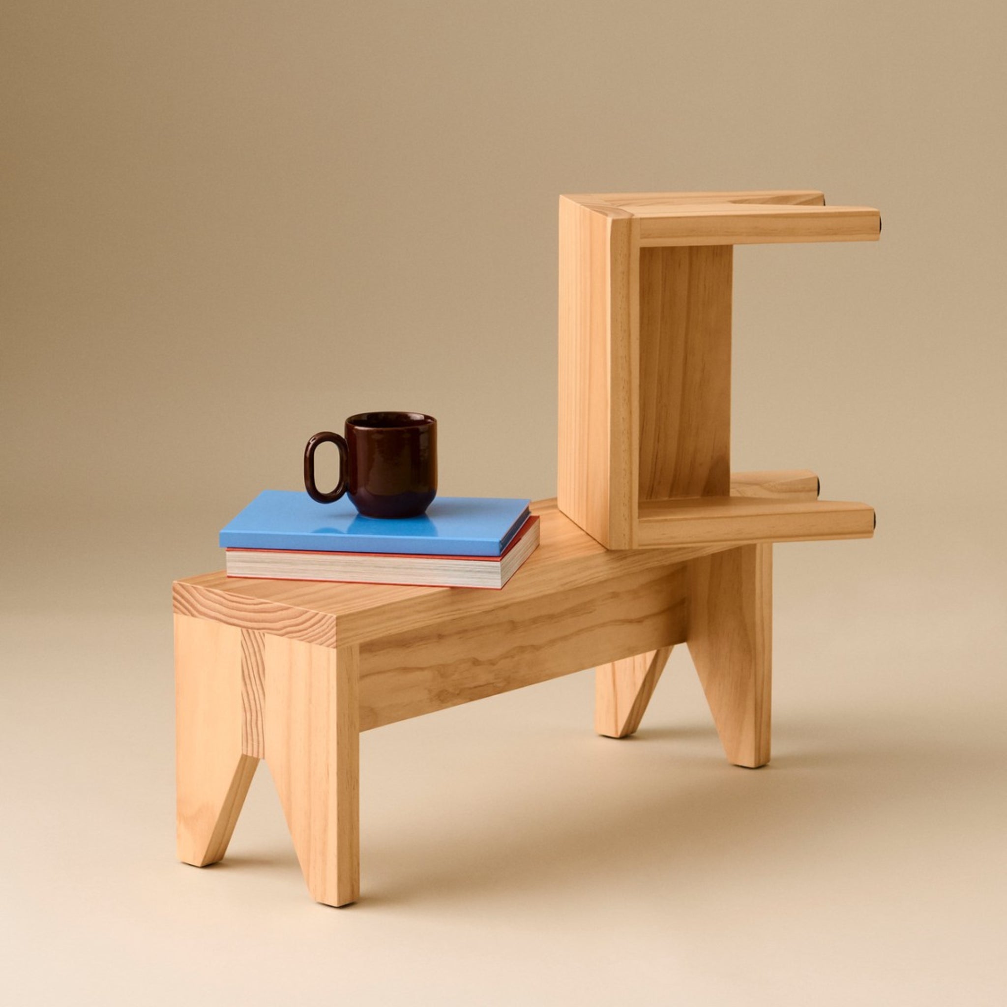 De Hay Manolito Kruk Bank Naturel Lang van Hay is een kleine, moderne houten bijzettafel met een L-vormige bovenste plank waarop een stapel boeken en een bruine mok staan, tegen een beige achtergrond.