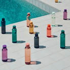 Hay Miz Waterfles Houtskool 720 ml van Hay staan rechtop op lichte tegels aan de rand van een zwembad, waarbij elke fles een schaduw werpt in het zonlicht.