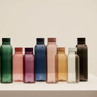 Een rij Hay Miz waterflessen in verschillende kleuren en maten op een witte sokkel, waaronder de Hay Miz Waterfles Karamel 540 ml.