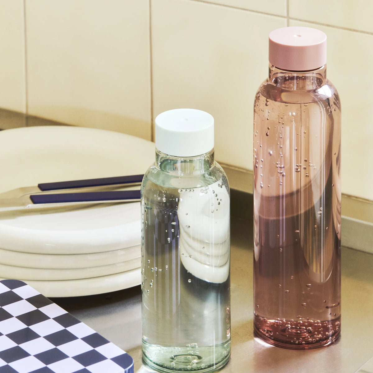 De Hay Miz Waterfles Mauve 720 ml met roze dop op een roestvrijstalen aanrecht, naast een heldere Miz fles, borden en een geblokte blikken doos.