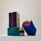 Drie Hay Multi Toilettas Medium Crème zakjes, met kleurrijke blokken en ritsen, zijn gestapeld op een wit oppervlak met het Hay logo zichtbaar op twee. De stoffen zakjes staan tegen een effen beige achtergrond.