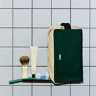 De Hay Multi Toilettas Medium Donkergroen van Hay staat op een betegeld rek met cosmetica zoals een tandenborstel, tandpasta, scheerkwast en scheermes ervoor - alles tegen een witte tegelwand.