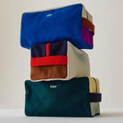 Drie rechthoekige HAY Multi Toilettas Medium Crème zijn gestapeld, elk gemaakt van biologisch katoen met een bijpassend handvat en rits. De merknaam "HAY" staat op de voorkant. De achtergrond is effen en licht.