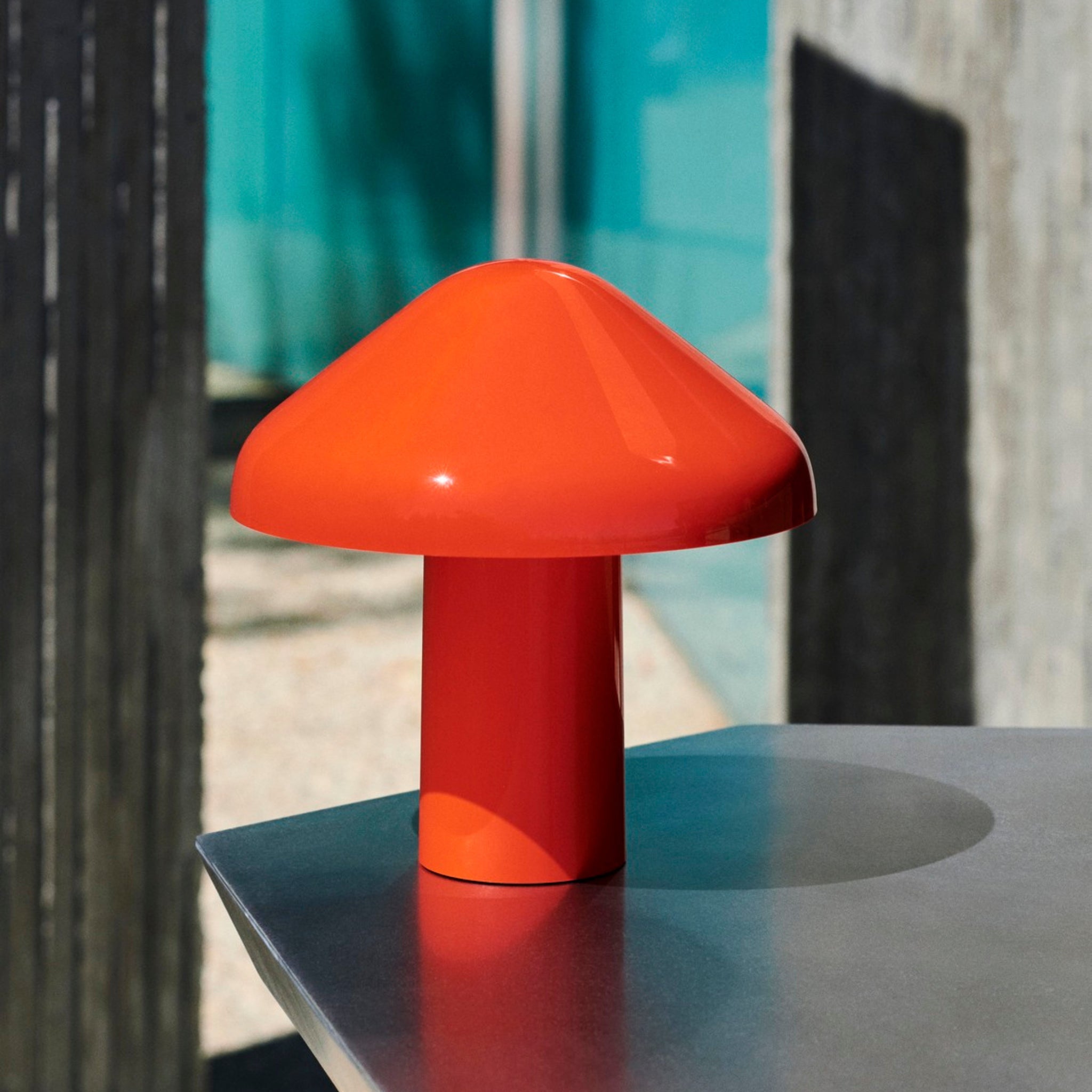 Een moderne Hay Pao Tafellamp LED Oplaadbaar Rood van Hay staat buiten op een grijs oppervlak. De paddestoelvorm en gedurfde rode kleur steken af tegen een vage achtergrond van groenblauw glas en beton.
