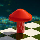 Een glanzend rode Hay Pao Tafellamp LED Oplaadbaar van Hay, geïnspireerd door Naoto Fukasawa, ligt op een groen en wit geruit oppervlak naast een zwembad met diepblauw water op de achtergrond.