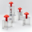 Vier metalen tafels met reflecterende oppervlakken, elk voorzien van een Hay Pao Tafellamp LED Oplaadbaar Rood van Hay. Ontworpen door Naoto Fukasawa, steken deze draadloze lampen af tegen een maagdelijk witte achtergrond, vrij van andere objecten.