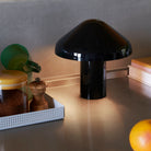 Een Hay Pao Tafellamp LED Oplaadbaar Zwart van Hay, ontworpen door Naoto Fukasawa, staat op een aanrecht naast een groen kopje, potje met geel deksel, pepermolen, boeken en een deels zichtbare sinaasappel op de voorgrond.