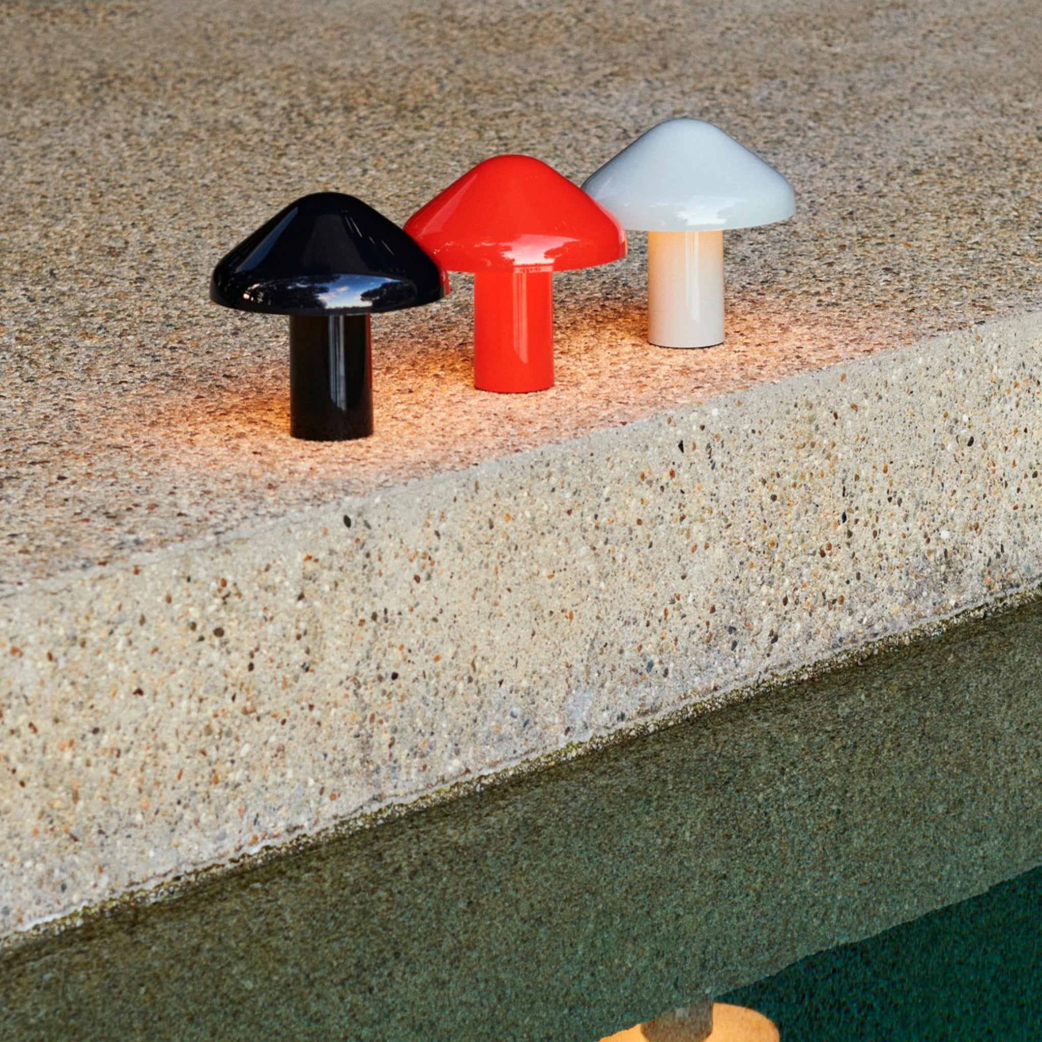 Drie kleine paddestoelvormige lampen in zwart, rood en wit - elk een Hay Pao Tafellamp LED Oplaadbaar Rood - schijnen op een gestructureerde steen boven groenachtig water. Hun elegante ontwerp is geïnspireerd op Naoto Fukasawa's Pao Portable Lamp.
