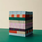 Een kubus van Hay Pattern Papieren Servetten Lichtroze Roos 33/33 van Hay, netjes gestapeld op een groen oppervlak tegen een beige achtergrond, laat levendige blauwe, rode, roze, groene en beige banden zien.