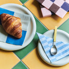 Een croissant ligt op een blauw en wit servet bovenop een bord, naast een ander bord met een lepel en Hay Pattern Papieren Servetten Grijs Blauw Stripe 33/33 van Hay, tegen geel en groen geruite tegels.