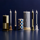 Vier Hay Patroon kaarsen in groen en blauw, elk met unieke geometrische patronen, staan rechtop op ronde voetstukken tegen een donkerblauwe achtergrond. Deze kaarsen van Hay dragen het Nordic Swan Ecolabel.