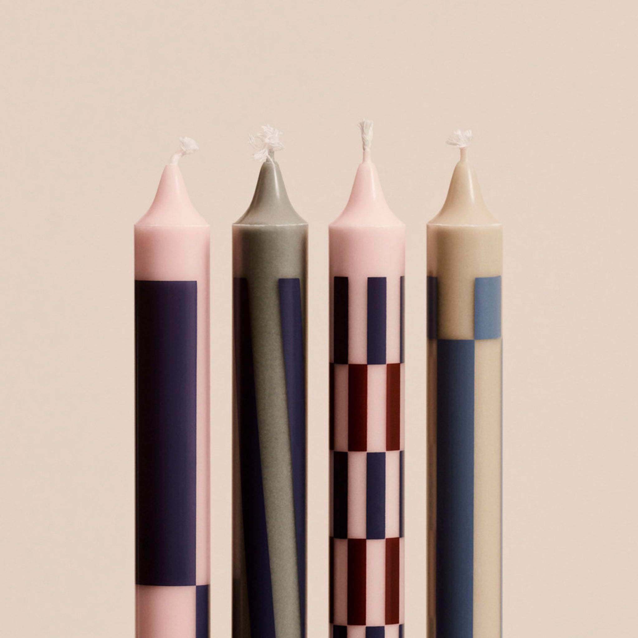 Vier hoge Hay Patroon Kaarsen Roze en Blauw (4 st.) van Hay, elk met een uniek geometrisch ontwerp in roze, blauw, grijs en bordeauxrood, staan naast elkaar. Gemaakt volgens de Nordic Swan Ecolabel normen; de lonten branden niet tegen een beige achtergrond.