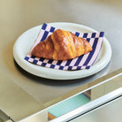 Een goudbruine croissant ligt op een Hay Pattern Papieren Servetten Lichtroze Donkerblauw Stripe 40/40 servet van Hay, bovenop een wit bord op een roestvrijstalen aanrecht.