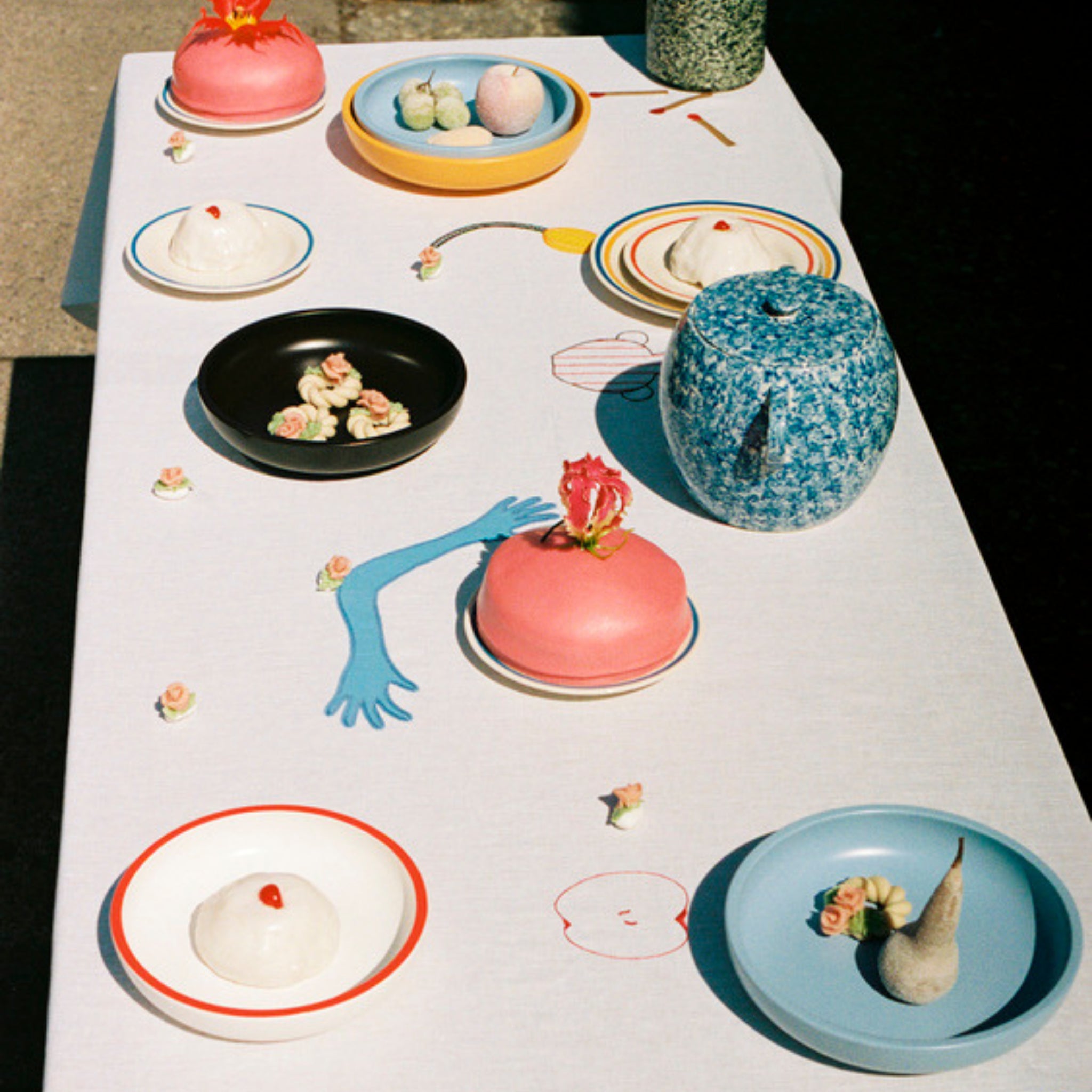 Op een grillige tafel staan pastelkleurige dessertachtige sculpturen, een blauwe HAY Laila Gohar theepot, de Hay Sobremesa Serveerschaal Geel D25 met een gehoornd object en speelse vormen die in het zonlicht op een wit tafelkleed zijn getekend.