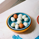 Een blauwe schaal met negen witte eieren en drie bruine eieren staat in een Hay Sobremesa Serveerschaal Geel D25 op een licht tafelkleed. Een gedeeltelijk glas en een blauwe handschoen in de buurt roepen de speelse stijl van HAY op.