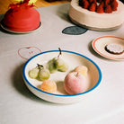 De Hay Sobremesa Serveerschaal Wit, Blauwe Rand D25 van Hay, gemaakt van ovenbestendig porselein, houdt gesuikerde druiven en een perzik op een tafel vol cake met een bord op een geborduurd tafelkleed.