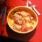 Een Hay Sobremesa Serveerschaal Geel D25 van Hay, gevuld met gesneden rode en gele tomaten, kerstomaatjes en gele bloemen, staat op een rood tafelkleed. Zilverwerk omringt de schaal terwijl het zonlicht zachte schaduwen over het tafereel werpt.