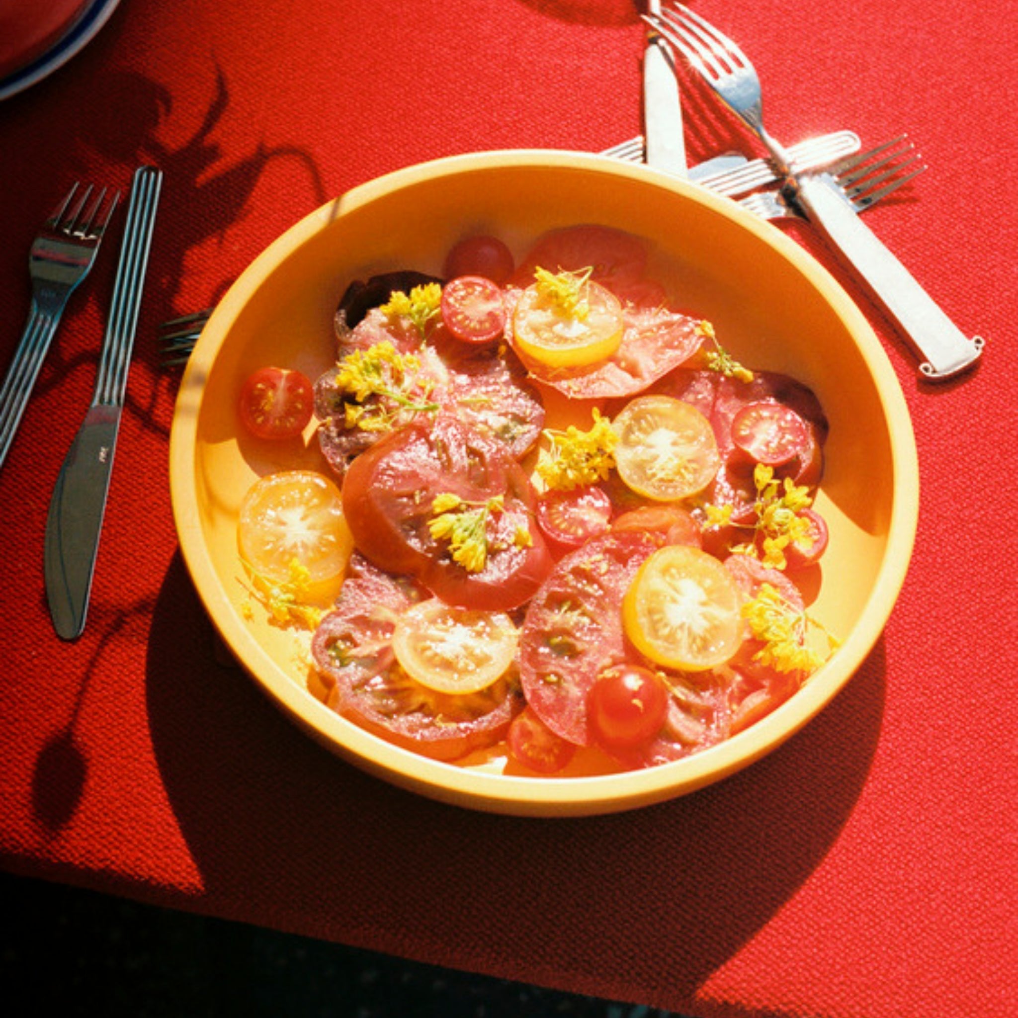 Een Hay Sobremesa Serveerschaal Geel D25 van Hay, gevuld met gesneden rode en gele tomaten, kerstomaatjes en gele bloemen, staat op een rood tafelkleed. Zilverwerk omringt de schaal terwijl het zonlicht zachte schaduwen over het tafereel werpt.