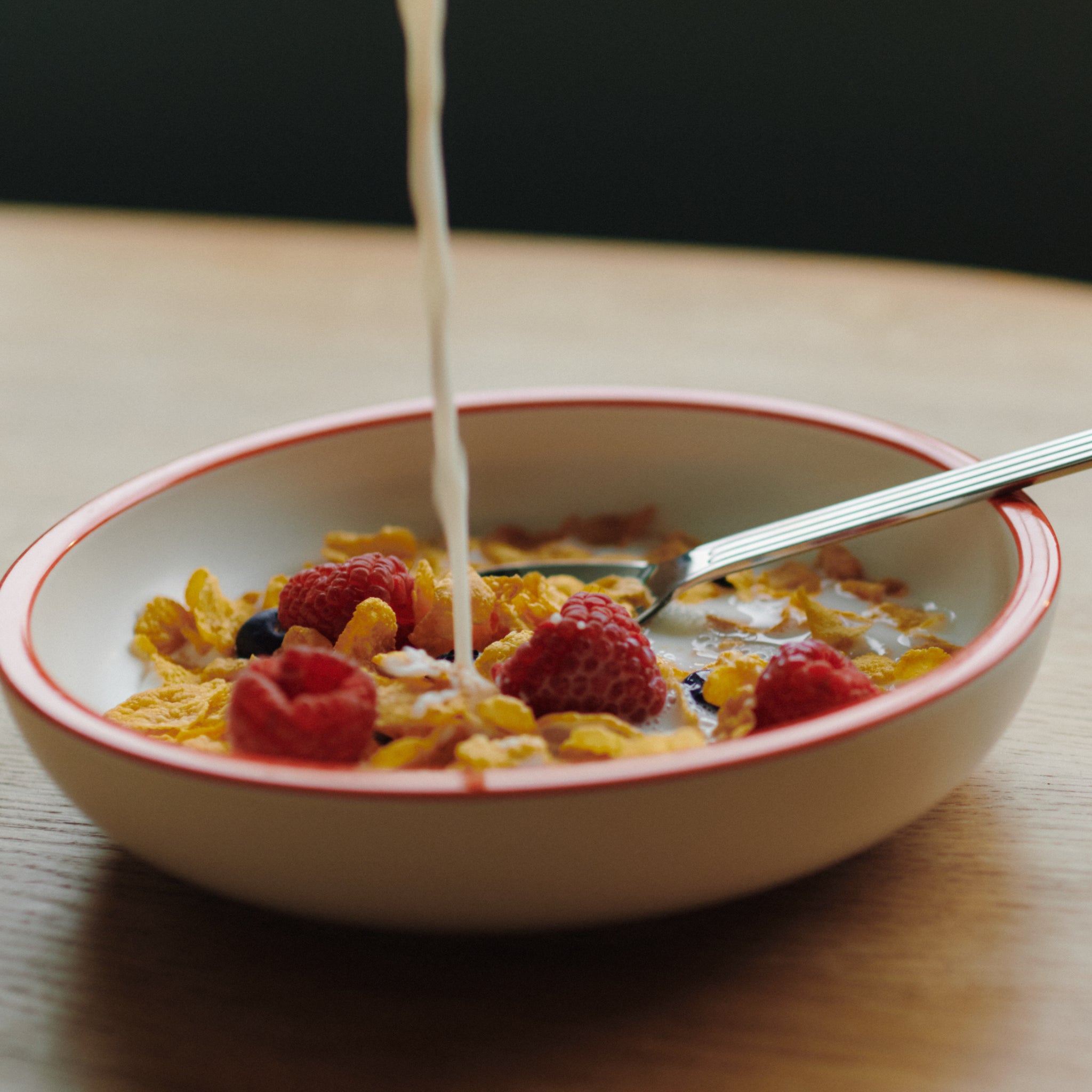 Een kom cornflakes met frambozen en bosbessen, melk wordt erbij gegoten. Een lepel rust in de Hay Sobremesa Serveerschaal Wit, Rode Rand D20 van Hay, die op een houten tafel staat.