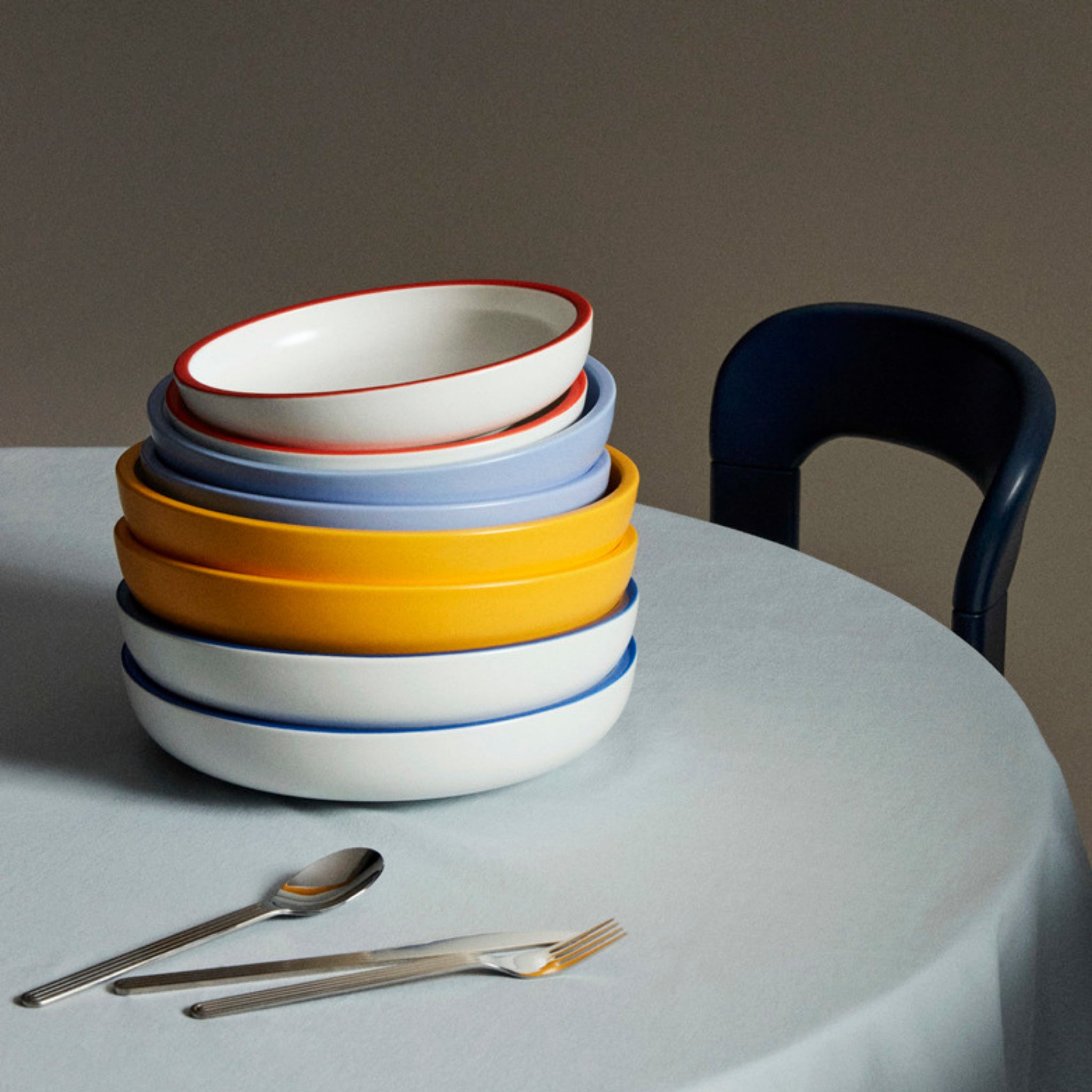 Een gele Hay Sobremesa Serveerschaal D25 serveerschotel zit op een ronde tafel met een lichtblauw tafelkleed, vergezeld van een vork en lepel; een deel van een donkere stoel verschijnt op de achtergrond.