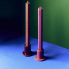 Twee Hay Tube Kandelaars met lange kaarsen op een blauw oppervlak tegen een groene muur, links de Hay Tube Kandelaar Kastanjebruin naast een bessenroze exemplaar.